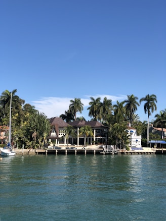 A luxurious waterfront house surrounded by tropical palm trees, with a clear blue sky overhead. The house is situated next to a calm waterway, with a small dock extending into the water. A white sailboat is moored nearby, adding to the serene and affluent ambiance.