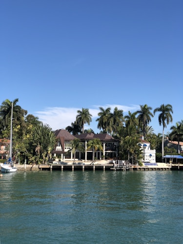 A luxurious waterfront house surrounded by tropical palm trees, with a clear blue sky overhead. The house is situated next to a calm waterway, with a small dock extending into the water. A white sailboat is moored nearby, adding to the serene and affluent ambiance.