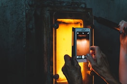 Hands holding a smartphone capturing an image of a glowing orange furnace or kiln. The scene is set in a dimly lit industrial environment, with the light from the furnace contrasting with the dark surroundings.