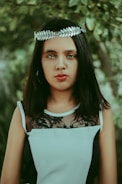 A person with long dark hair is wearing a silver laurel leaf headband and a sleeveless black and white dress with lace detailing. The background features lush green foliage, adding a natural and serene setting.