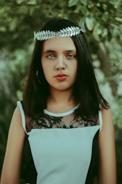 A person with long dark hair is wearing a silver laurel leaf headband and a sleeveless black and white dress with lace detailing. The background features lush green foliage, adding a natural and serene setting.