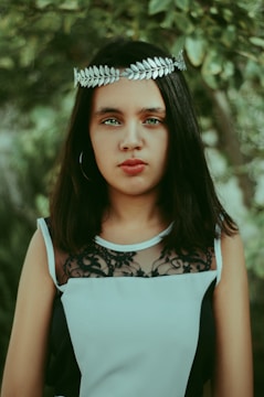 A person with long dark hair is wearing a silver laurel leaf headband and a sleeveless black and white dress with lace detailing. The background features lush green foliage, adding a natural and serene setting.