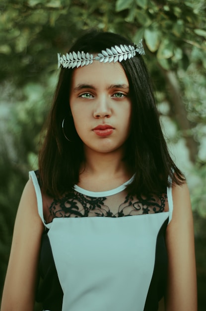 A person with long dark hair is wearing a silver laurel leaf headband and a sleeveless black and white dress with lace detailing. The background features lush green foliage, adding a natural and serene setting.
