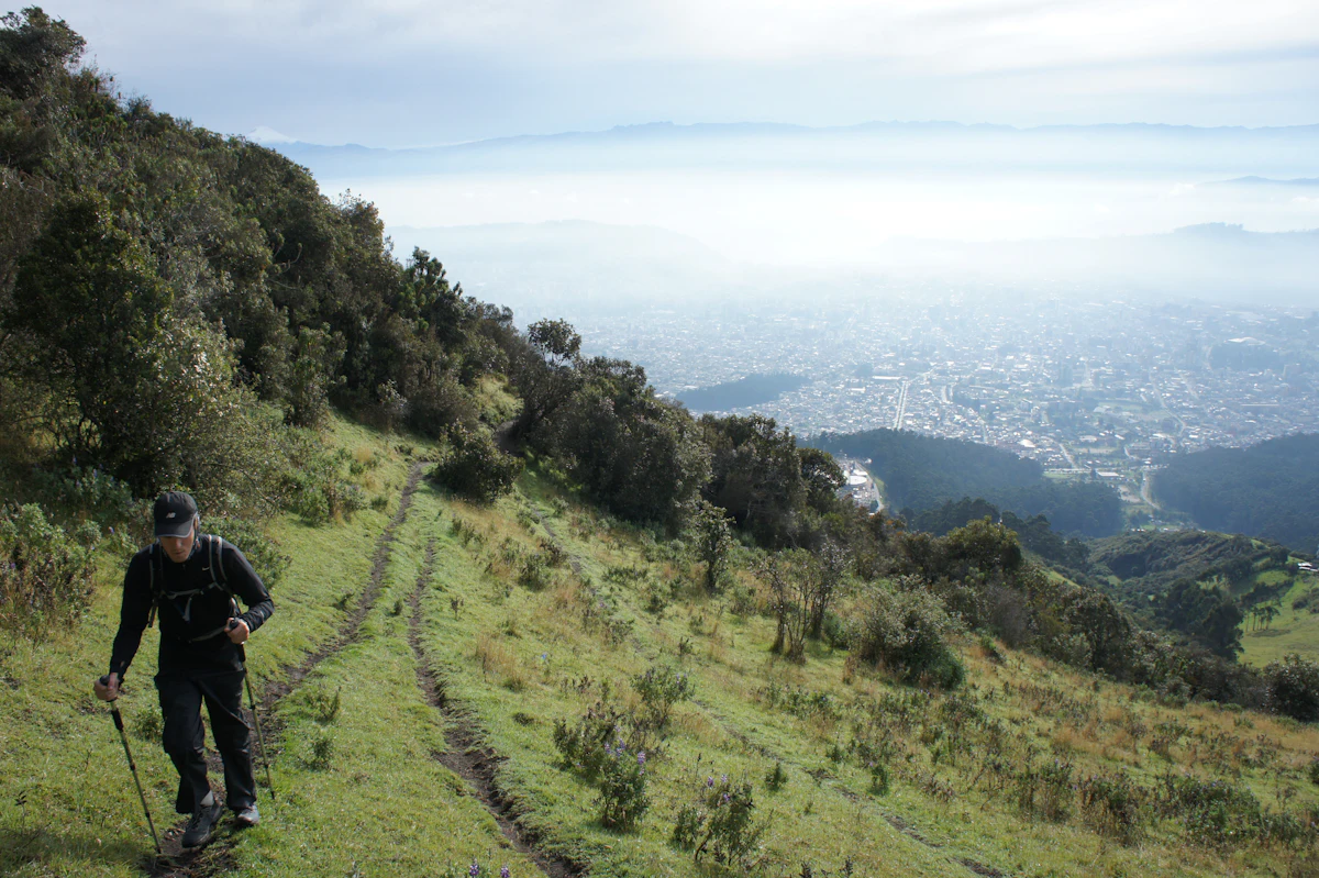A hiker with a trekking pole on a grassy path near the TelefériQo, on the way up Rucu Pichincha, with Quito below