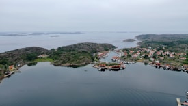 The image depicts a coastal landscape with a serene body of water surrounded by rocky hills and a picturesque village. The village features numerous small houses with red roofs, interspersed with green vegetation. Several boats are moored along the shoreline, with a few docks extending into the water. The sky is overcast, contributing to a calm and peaceful atmosphere.