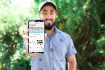 A person stands outdoors, holding a smartphone prominently towards the camera. The phone's screen displays a website with various items including a tandem bike and a portable propane fireplace. The background is filled with lush green foliage, creating a natural setting.