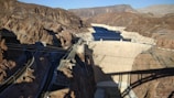 A large concrete dam set amidst a rocky desert landscape, with a reservoir of dark blue water behind it. Power lines and roads are visible in the foreground, along with rugged terrain featuring reddish-brown mountains. The shadow of a bridge partially covers the dam.