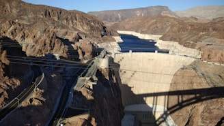 A large concrete dam set amidst a rocky desert landscape, with a reservoir of dark blue water behind it. Power lines and roads are visible in the foreground, along with rugged terrain featuring reddish-brown mountains. The shadow of a bridge partially covers the dam.