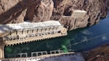 A large hydroelectric power station is constructed amidst rocky cliffs with a river flowing beside it. The station features multiple generators and complex electrical infrastructure on the roof. The building is integrated into the steep natural terrain, and the water appears to be a deep blue-green color.