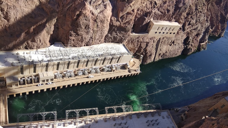A large hydroelectric power station is constructed amidst rocky cliffs with a river flowing beside it. The station features multiple generators and complex electrical infrastructure on the roof. The building is integrated into the steep natural terrain, and the water appears to be a deep blue-green color.