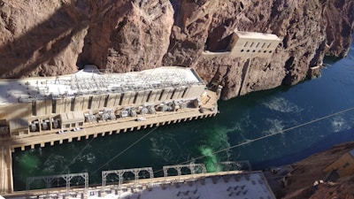 A large hydroelectric power station is constructed amidst rocky cliffs with a river flowing beside it. The station features multiple generators and complex electrical infrastructure on the roof. The building is integrated into the steep natural terrain, and the water appears to be a deep blue-green color.