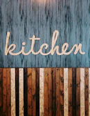 A stylized wooden sign spelling out the word 'kitchen' is mounted on a vertically striped background. The top half of the image features a blue and white striped pattern, while the lower half consists of alternating wooden planks in various shades of brown. The design combines a rustic and modern aesthetic.