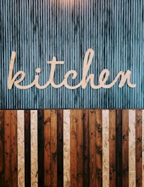 A stylized wooden sign spelling out the word 'kitchen' is mounted on a vertically striped background. The top half of the image features a blue and white striped pattern, while the lower half consists of alternating wooden planks in various shades of brown. The design combines a rustic and modern aesthetic.