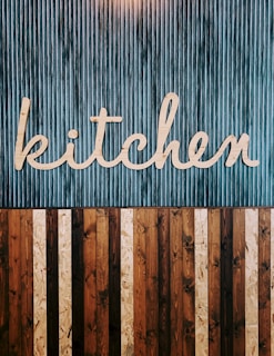A stylized wooden sign spelling out the word 'kitchen' is mounted on a vertically striped background. The top half of the image features a blue and white striped pattern, while the lower half consists of alternating wooden planks in various shades of brown. The design combines a rustic and modern aesthetic.