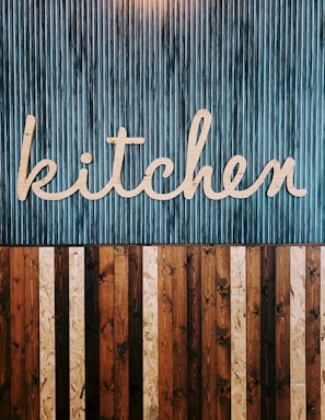 A stylized wooden sign spelling out the word 'kitchen' is mounted on a vertically striped background. The top half of the image features a blue and white striped pattern, while the lower half consists of alternating wooden planks in various shades of brown. The design combines a rustic and modern aesthetic.