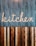 A stylized wooden sign spelling out the word 'kitchen' is mounted on a vertically striped background. The top half of the image features a blue and white striped pattern, while the lower half consists of alternating wooden planks in various shades of brown. The design combines a rustic and modern aesthetic.
