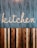 A stylized wooden sign spelling out the word 'kitchen' is mounted on a vertically striped background. The top half of the image features a blue and white striped pattern, while the lower half consists of alternating wooden planks in various shades of brown. The design combines a rustic and modern aesthetic.