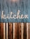 A stylized wooden sign spelling out the word 'kitchen' is mounted on a vertically striped background. The top half of the image features a blue and white striped pattern, while the lower half consists of alternating wooden planks in various shades of brown. The design combines a rustic and modern aesthetic.