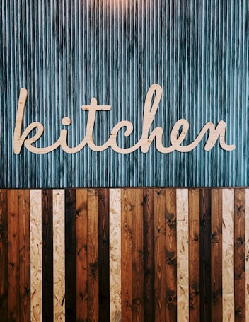 A stylized wooden sign spelling out the word 'kitchen' is mounted on a vertically striped background. The top half of the image features a blue and white striped pattern, while the lower half consists of alternating wooden planks in various shades of brown. The design combines a rustic and modern aesthetic.