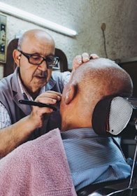 An elderly man wearing glasses is carefully shaving the face of a bald man seated in a barber chair, with a towel draped around his shoulders. The setting appears to be a traditional barbershop, characterized by a vintage and cozy atmosphere.