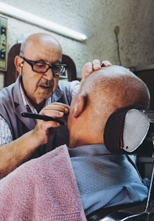 An elderly man wearing glasses is carefully shaving the face of a bald man seated in a barber chair, with a towel draped around his shoulders. The setting appears to be a traditional barbershop, characterized by a vintage and cozy atmosphere.