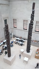 Two large, intricately carved totem poles are positioned indoors within a spacious room. Several people are seated at tables, some engaged in conversation or using laptops. The walls are high and feature large windows, allowing natural light to illuminate the area.
