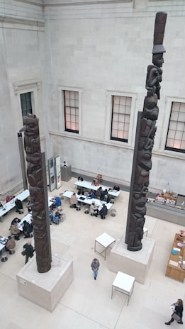 Two large, intricately carved totem poles are positioned indoors within a spacious room. Several people are seated at tables, some engaged in conversation or using laptops. The walls are high and feature large windows, allowing natural light to illuminate the area.