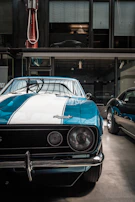 A vintage car with a blue and white paint job, prominently featuring a classic design with a chrome bumper and round headlights. The setting appears to be indoors, possibly in a museum or showroom, as suggested by the industrial lighting and the modern car partially visible through the windows above.