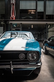 A vintage car with a blue and white paint job, prominently featuring a classic design with a chrome bumper and round headlights. The setting appears to be indoors, possibly in a museum or showroom, as suggested by the industrial lighting and the modern car partially visible through the windows above.