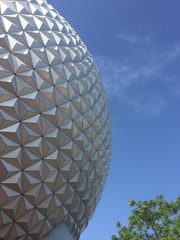 A large geodesic sphere with a geometric pattern against a clear blue sky. Some green tree branches are visible in the lower right corner.