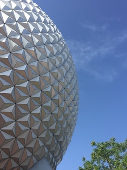 A large geodesic sphere with a geometric pattern against a clear blue sky. Some green tree branches are visible in the lower right corner.