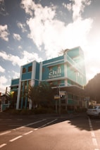 A vibrant turquoise building with the words 'chill backpackers' is bathed in warm sunlight. The structure is situated at a street corner, surrounded by palm trees and various greenery. The sky is blue with scattered clouds, and a car is parked nearby.