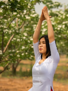 A person is engaged in a yoga pose with hands pressed together overhead in an outdoor setting. The background features lush green trees with white blossoms, suggesting a spring or early summer season.