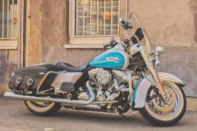 A classic Harley-Davidson motorcycle is parked on a street with a vintage design and an eye-catching turquoise and chrome color scheme. The bike features chrome detailing, leather saddlebags, and a comfortable seat. It is positioned in front of a weathered building with barred windows and faded walls.