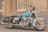A classic Harley-Davidson motorcycle is parked on a street with a vintage design and an eye-catching turquoise and chrome color scheme. The bike features chrome detailing, leather saddlebags, and a comfortable seat. It is positioned in front of a weathered building with barred windows and faded walls.