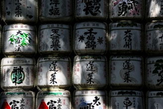 Rows of traditional Japanese sake barrels with intricate calligraphy and designs, stacked closely together. Shadows and light play across the surface, highlighting the barrels' textures and decorations.