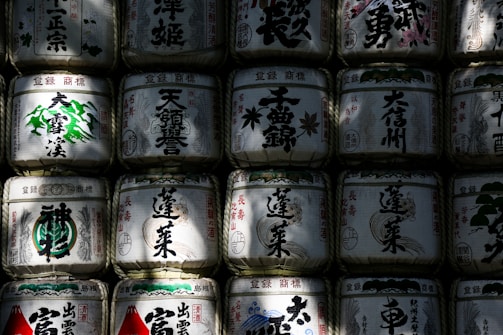 Rows of traditional Japanese sake barrels with intricate calligraphy and designs, stacked closely together. Shadows and light play across the surface, highlighting the barrels' textures and decorations.