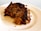 A slice of cake featuring a rich, crumbly texture with visible chunks of nuts and dried fruits. The cake appears moist with a slightly caramelized, dark brown crust, resting on a white plate.
