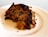 A slice of cake featuring a rich, crumbly texture with visible chunks of nuts and dried fruits. The cake appears moist with a slightly caramelized, dark brown crust, resting on a white plate.