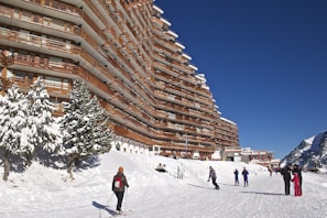 A large, multi-story building with wooden balconies lines a snow-covered ski slope. Skiers and snowboarders are visible enjoying the winter activity. Evergreen trees are coated with snow, and the sky is a clear, deep blue.