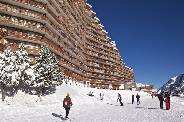 A large, multi-story building with wooden balconies lines a snow-covered ski slope. Skiers and snowboarders are visible enjoying the winter activity. Evergreen trees are coated with snow, and the sky is a clear, deep blue.