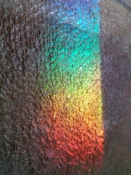 A close-up view of a textured surface with a vibrant spectrum of colors ranging from blue to red, resembling a rainbow effect. The surface has a rough, fibrous appearance, enhancing the iridescent quality of the colors.