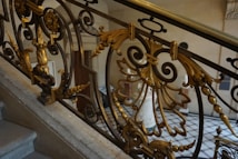 A detailed wrought iron railing is adorned with ornate, golden embellishments and intricate floral designs. The balustrade is part of a staircase in an elegantly decorated interior space with marble steps and classical architecture.