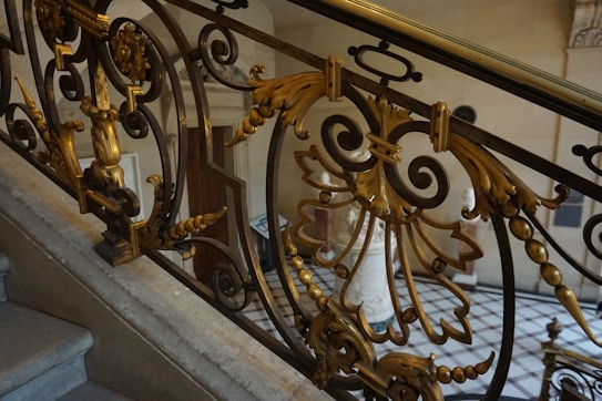 A detailed wrought iron railing is adorned with ornate, golden embellishments and intricate floral designs. The balustrade is part of a staircase in an elegantly decorated interior space with marble steps and classical architecture.
