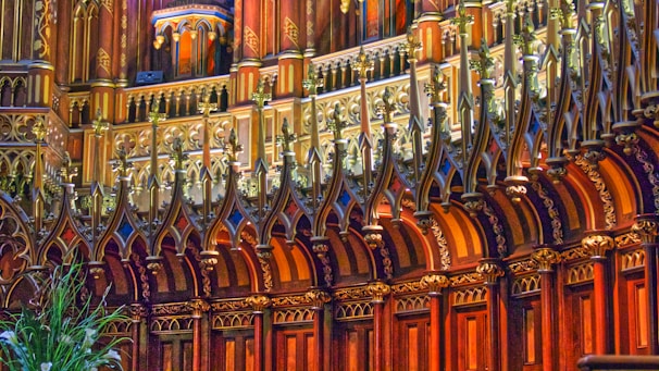 An ornate section of a building featuring intricate wooden carvings and gilded decorations. The design includes geometric and floral patterns, highlighted by ambient lighting. The architecture is Gothic-inspired with pointed arches and richly detailed moldings.