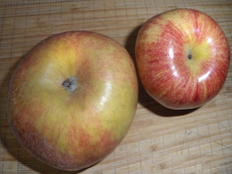 Two apples are placed on a wooden surface. The apple on the left is larger, with a mutted green and red hue, while the one on the right is smaller, with a more vibrant red and yellow coloration. The wooden surface features visible grid lines and measurement markings.
