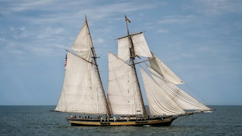 A two-masted schooner with cream sails on a calm sea