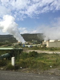 A large industrial facility with several pipes and structures emitting steam, set against a backdrop of green hills under a partly cloudy sky.