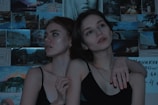 Two young individuals are posed closely together against a backdrop of various photographs and artwork. The lighting is dim and moody, adding a dramatic effect to their expressions which appear contemplative and introspective.