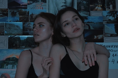 Two young individuals are posed closely together against a backdrop of various photographs and artwork. The lighting is dim and moody, adding a dramatic effect to their expressions which appear contemplative and introspective.